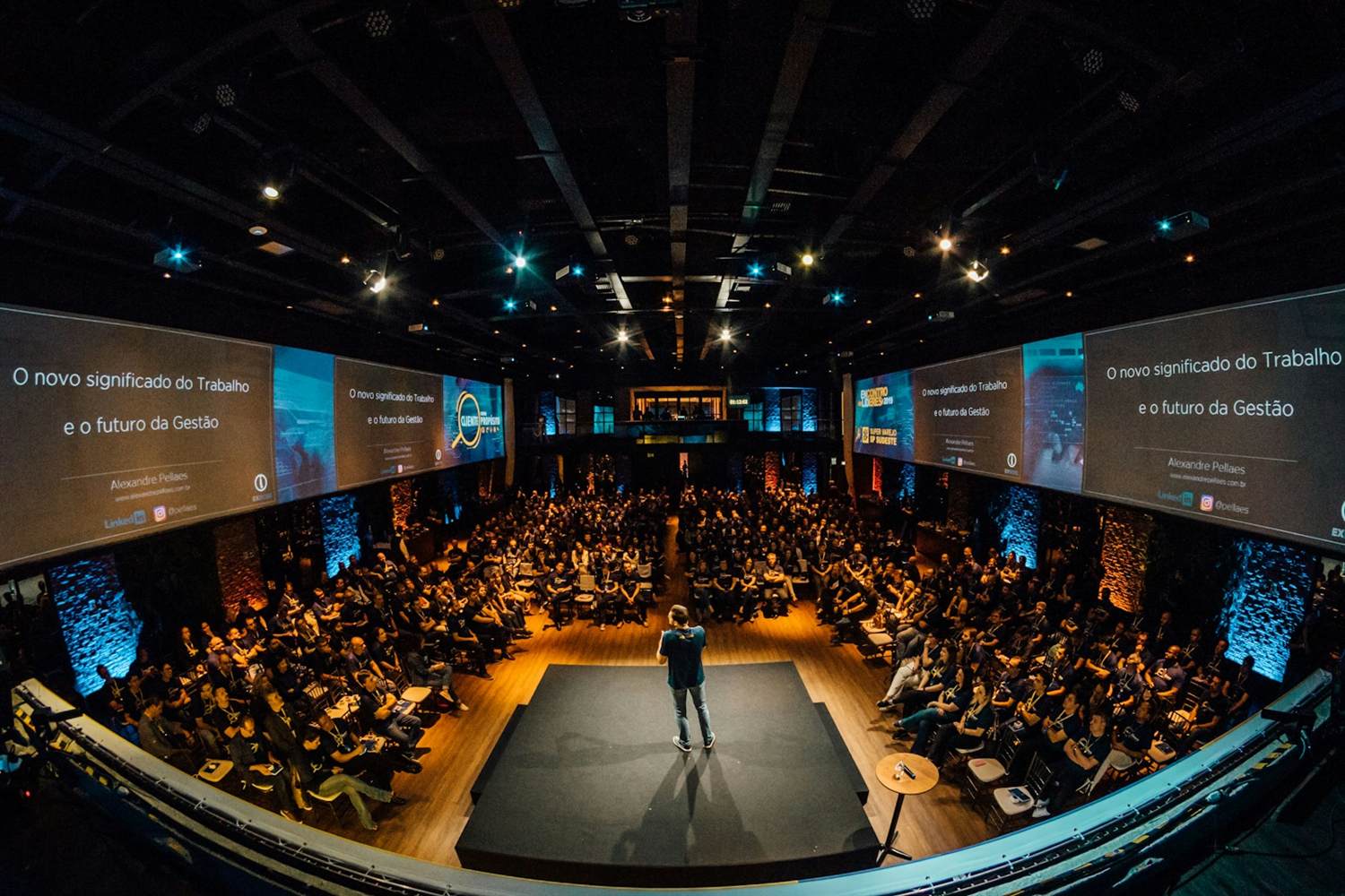 A speaker on a stage in front of a packed conference audience under wide screens