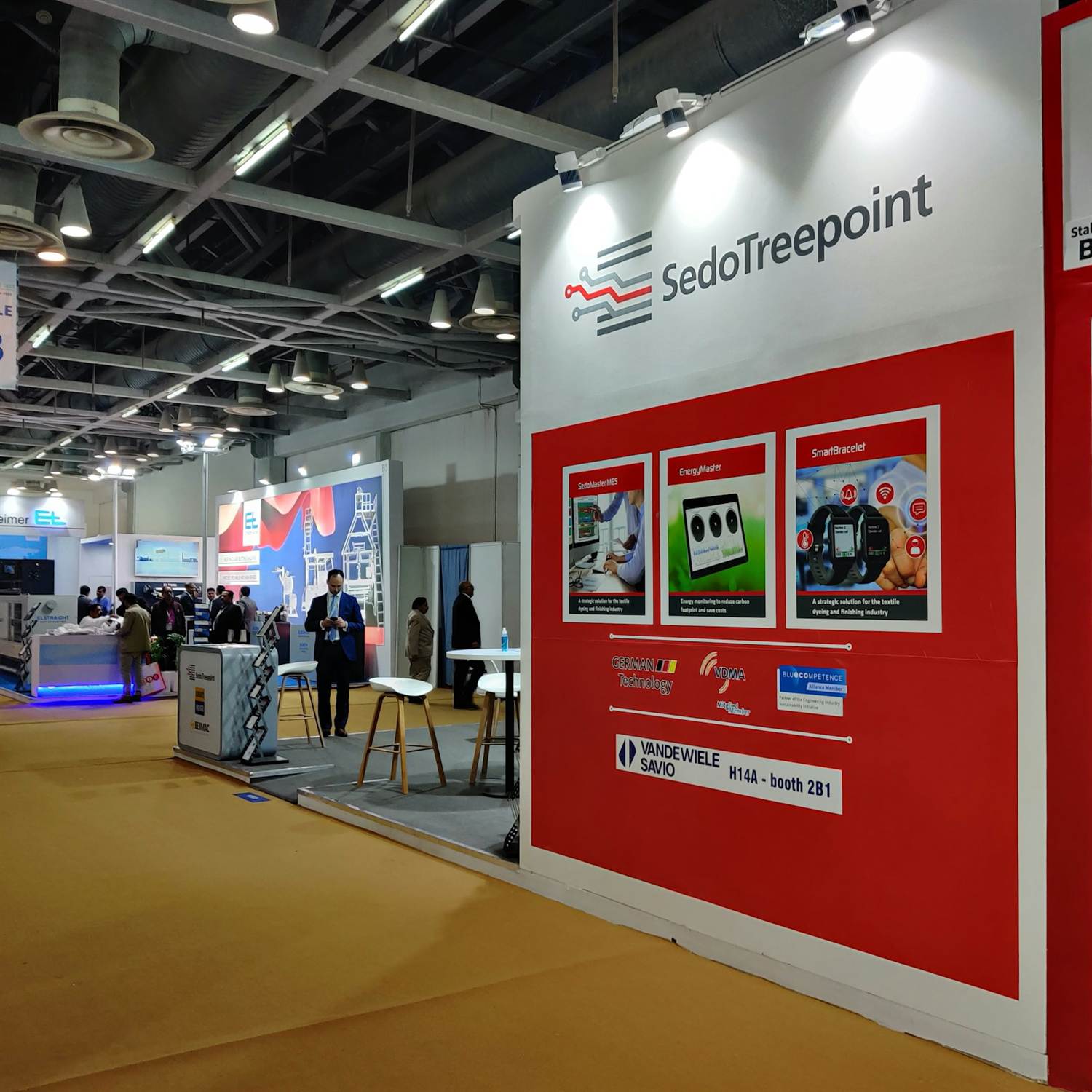 Red and white inline trade show booth with product panels, stools and a meeting table on a show floor