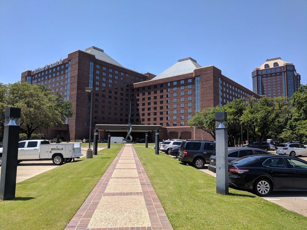 The brick Atrium wings of the Hilton Anatole with the hotel name on the roofline and the glass-topped Tower behind, seen from the front walk and parking lot
