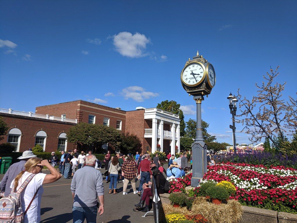 Fairgoers walking past a brick colonial-style building with white columns, a post clock and red and white flower beds on the Big E grounds in West Springfield