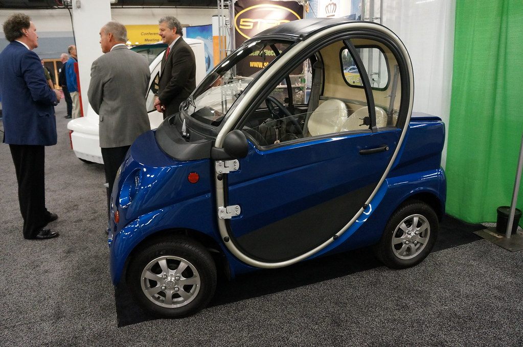 A small blue two-seat electric car on display on the carpeted exhibit floor of the Greenville Convention Center, then the TD Convention Center, with visitors talking between booths behind it