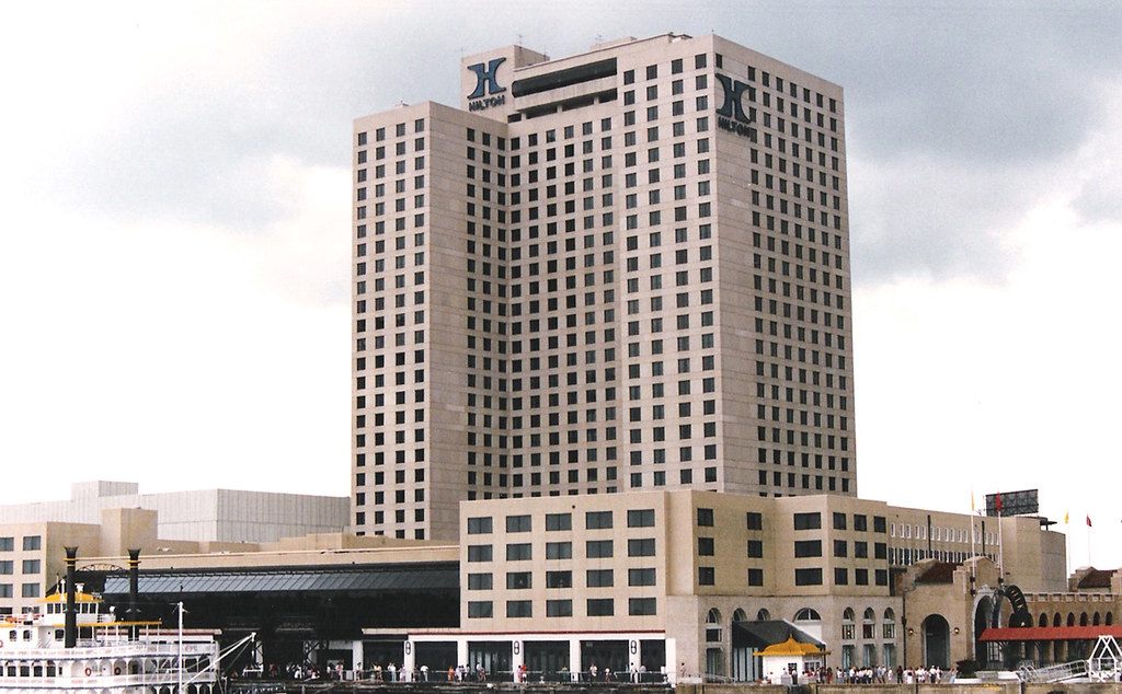 The Hilton New Orleans Riverside tower with Hilton logos at the top, rising behind its lower riverfront wing, with a paddlewheeler docked at left