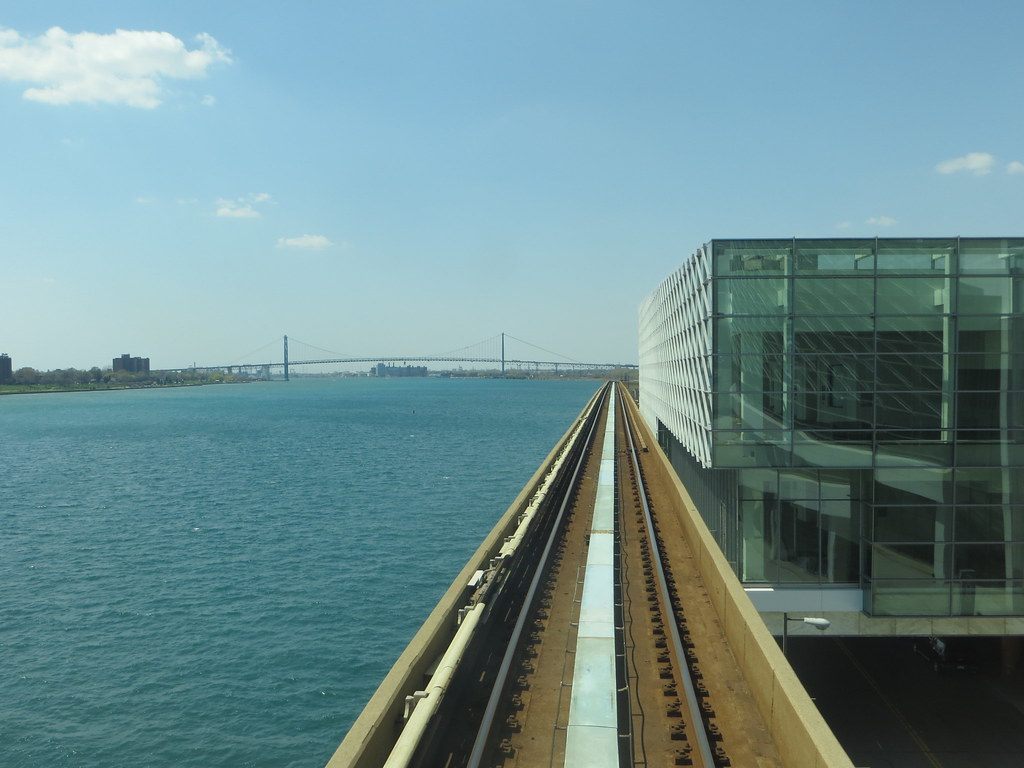 The Detroit People Mover track running past the glass riverfront atrium of Huntington Place, with the Detroit River and the Ambassador Bridge beyond