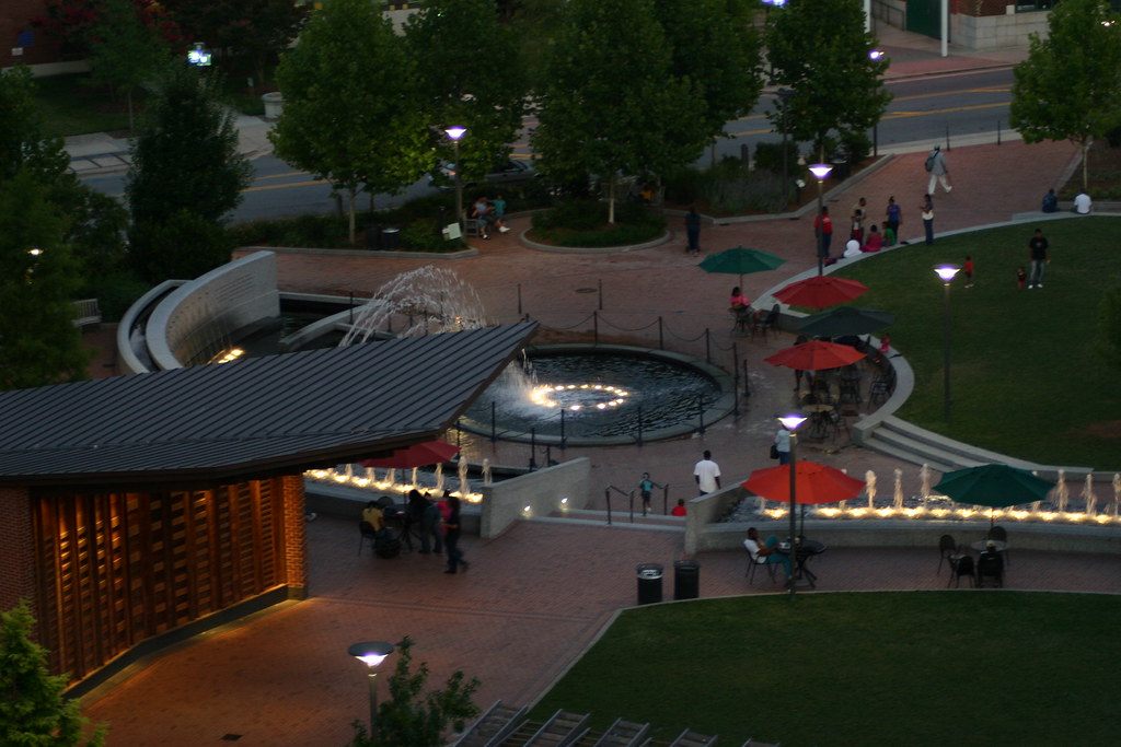 Diners under red and green umbrellas around the lit fountain in Center City Park, downtown Greensboro, at dusk