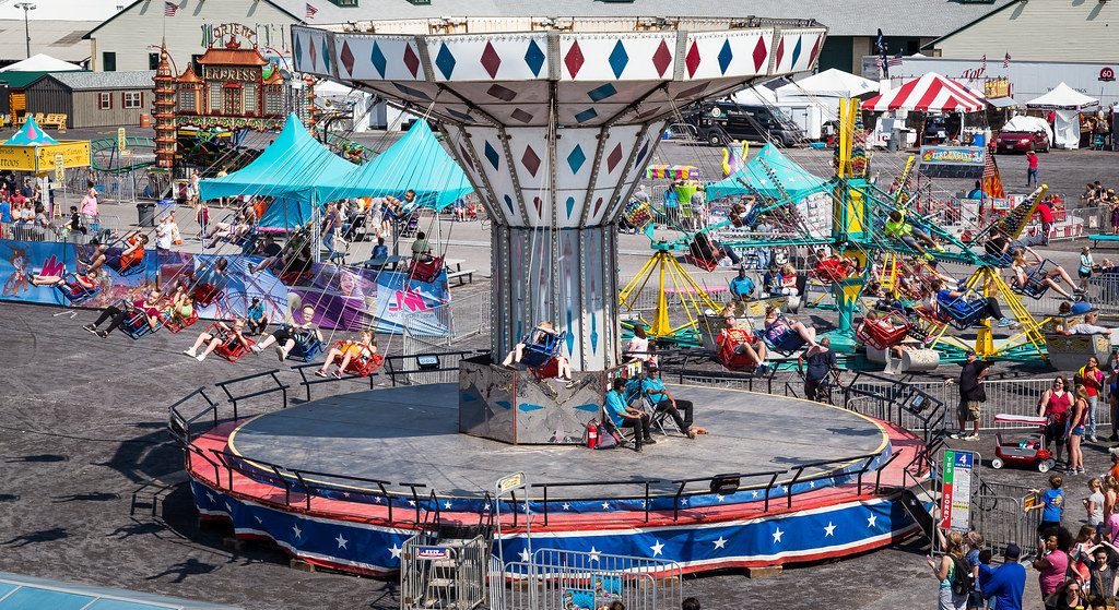 Swing ride, kiddie rides and striped tents on the midway of the New York State Fairgrounds during the Great New York State Fair, with crowds walking between them