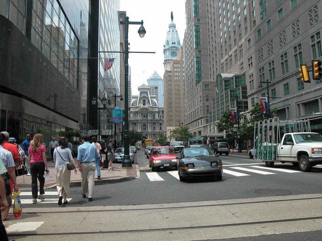 Pedestrians crossing Market Street at 12th Street, with office and hotel towers on both sides and City Hall's clock tower at the end of the street