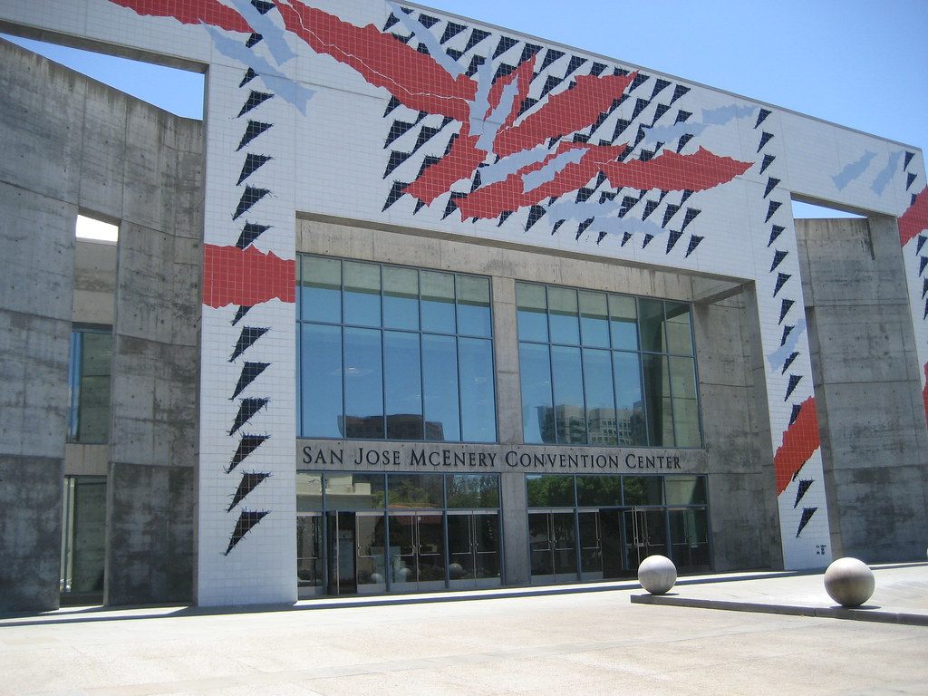 The glass main entrance of the San Jose McEnery Convention Center, framed by concrete walls and a red, white and black tile mural