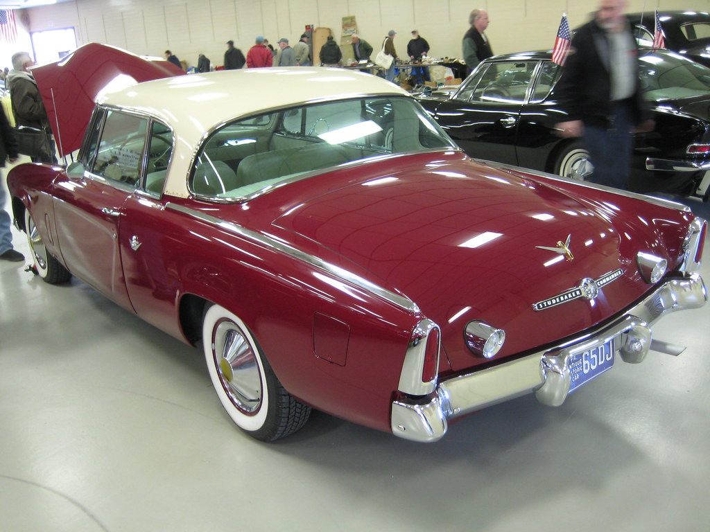 A maroon and cream Studebaker coupe parked on the concrete floor of a York fairgrounds exhibit hall during a Studebaker Drivers Club meet, with visitors browsing swap-meet tables behind it