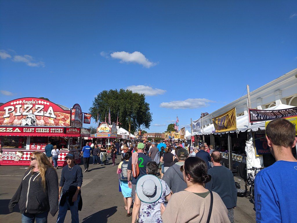 Crowds walking down a row of food stands and vendor tents on the Big E grounds under a clear blue sky