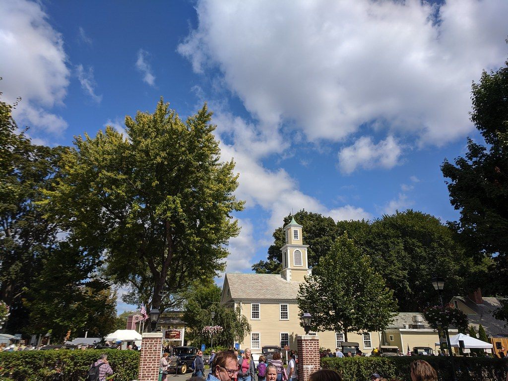 A white clapboard building with a steeple behind tall trees and a brick gateway, with fairgoers in the foreground