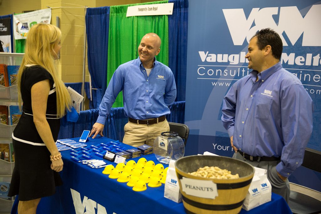 Two exhibitors in matching blue shirts talking with a visitor across a table of giveaways at a pipe-and-drape booth, North Charleston Business Expo