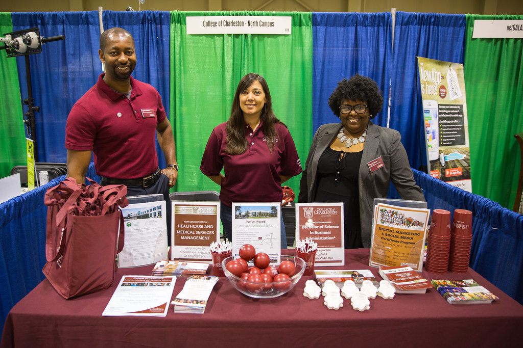 Three college staff members standing behind a maroon-draped table of brochures and giveaways at a booth, North Charleston Business Expo