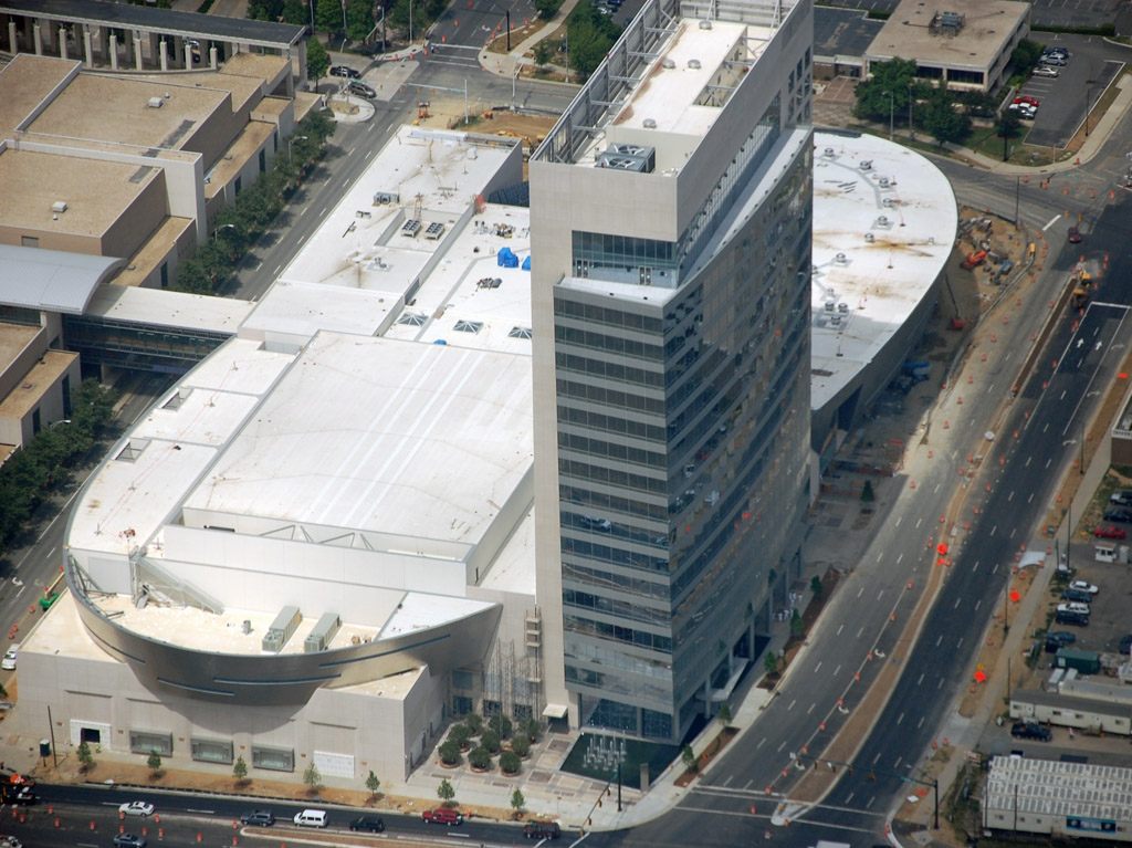 Aerial view of the NASCAR Hall of Fame office tower and curved ballroom roof under construction, with the low flat roofs of neighboring Uptown buildings