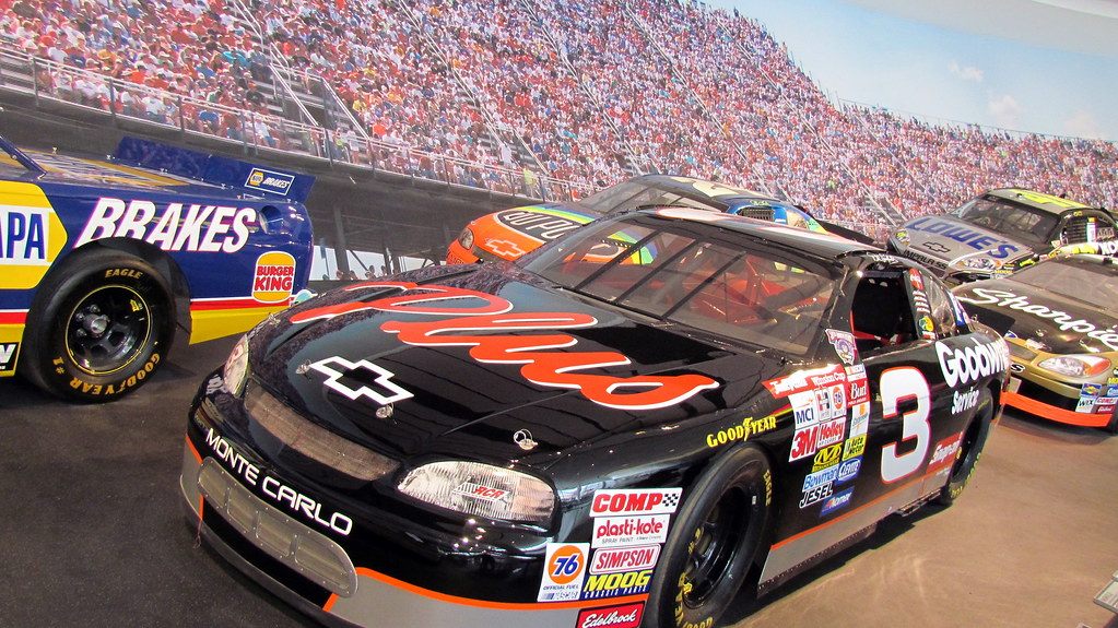 Stock cars, including a black No. 3 Chevrolet Monte Carlo, on display in front of a grandstand mural inside the NASCAR Hall of Fame
