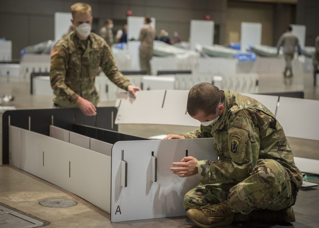 Two Connecticut National Guard members assembling white partition panels on the bare concrete exhibit hall floor, with a floor utility box in the foreground