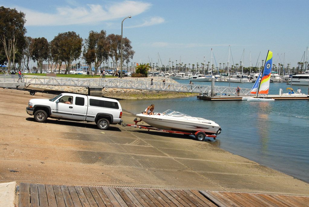 A pickup truck backing a small motorboat on a trailer down a concrete launch ramp beside a marina full of sailboats