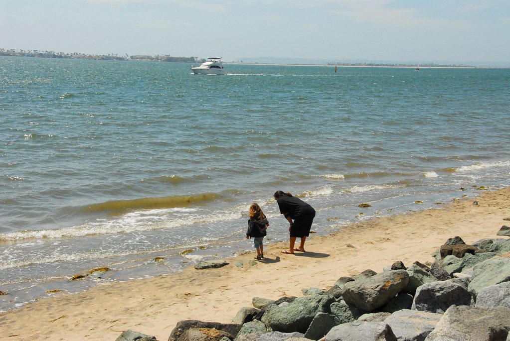 An adult and a small child exploring a sandy shoreline by rocks as a motor yacht passes on San Diego Bay