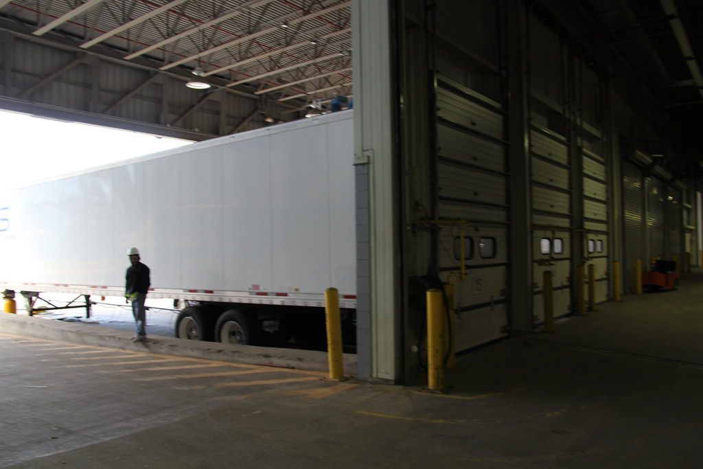 A white semitrailer backed into the covered truck dock for Halls A–D, beside a row of numbered dock doors