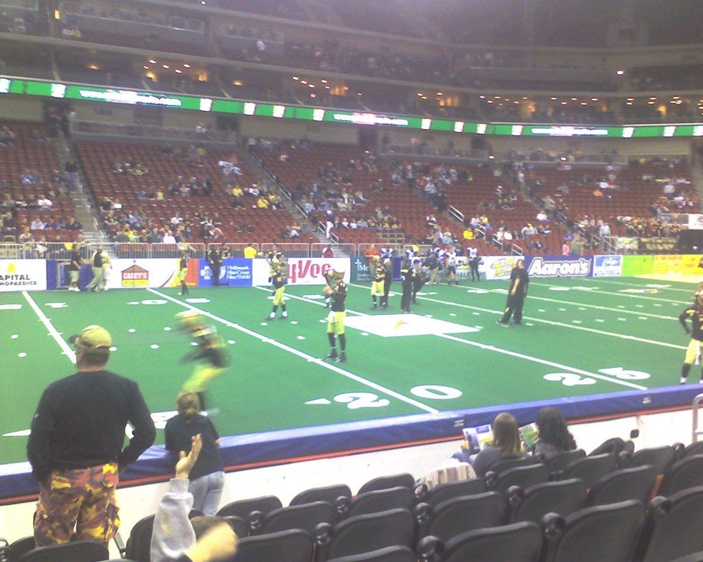 The arena bowl set up for an indoor football game, with green turf, players on the field and fans in the lower seats