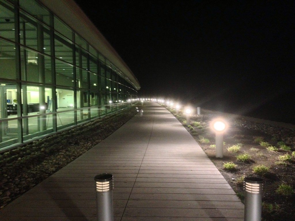 A lit boardwalk running at night along the glass wall of the Erie convention center, with bollard lights on one side
