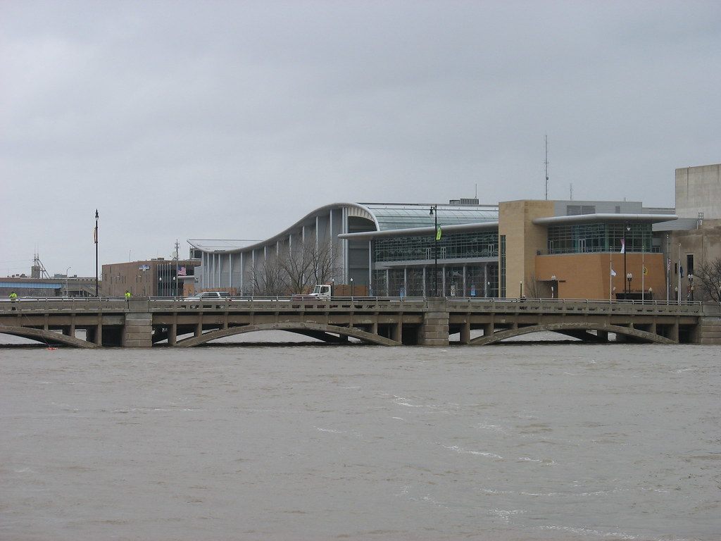 DeVos Place seen across the Grand River and an arched road bridge, with the long curved roof of the exhibit hall and the glass Grand Gallery behind it