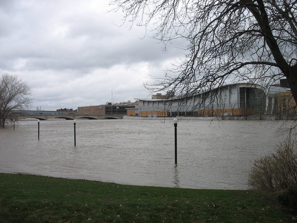 The long curved roof of DeVos Place's exhibit hall seen across the swollen Grand River from Ah-Nab-Awen Park, with bare trees in the foreground