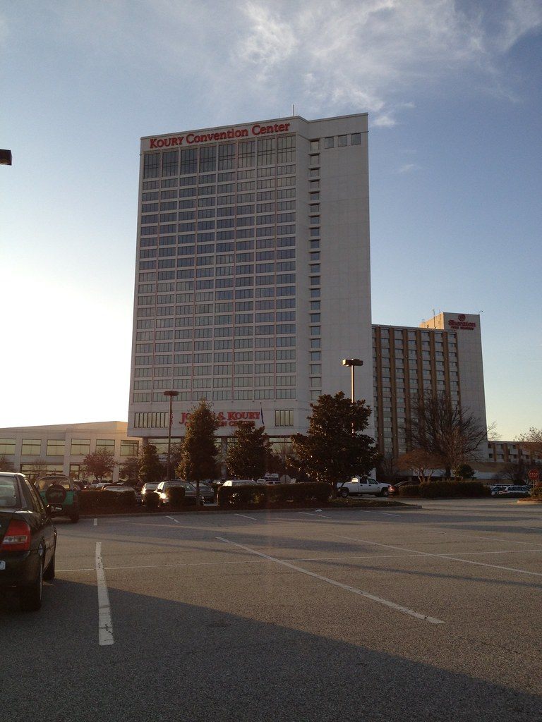 The white high-rise tower of the Sheraton Greensboro with Koury Convention Center lettered across the top, seen across a parking lot at dusk