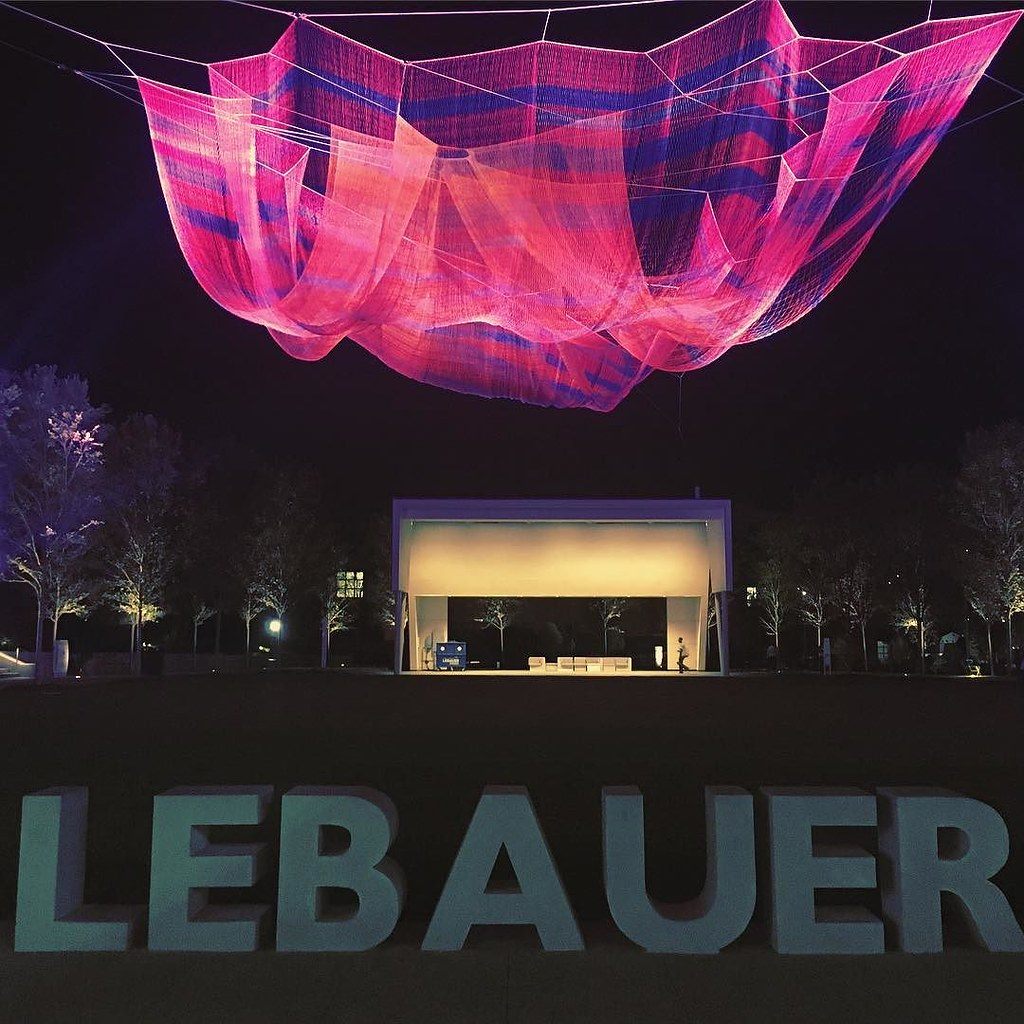 A pink and purple suspended net sculpture glowing over the lawn and stage of LeBauer Park at night