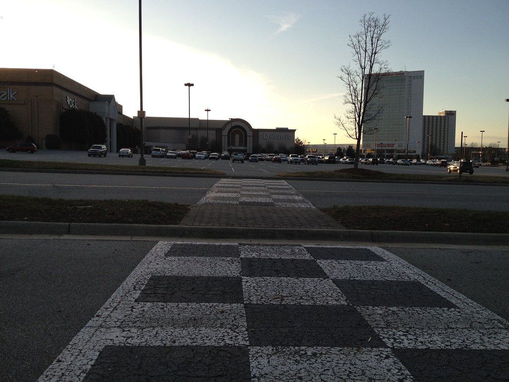 Four Seasons Town Centre’s Belk and Dillard’s stores across the parking lot at sunset, with the Sheraton Greensboro tower over the Koury Convention Center at right