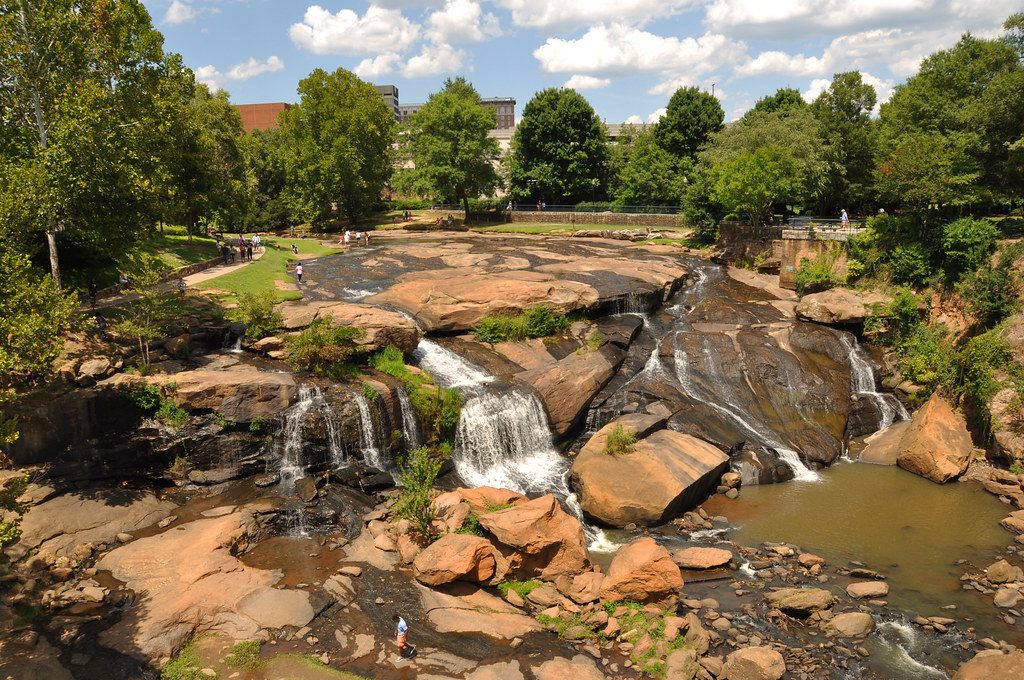 Reedy River Falls tumbling over granite ledges in Falls Park, with walkers on the lawns and downtown Greenville buildings behind the trees