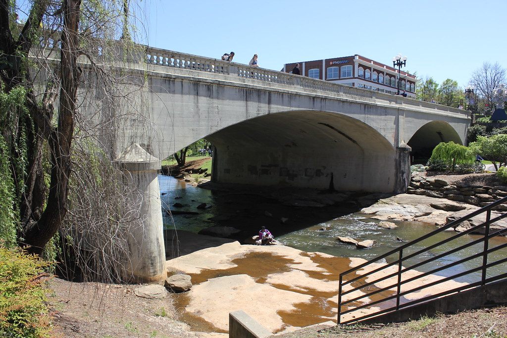 The arched concrete Main Street Bridge over the Reedy River in downtown Greenville, with people on the rocks below