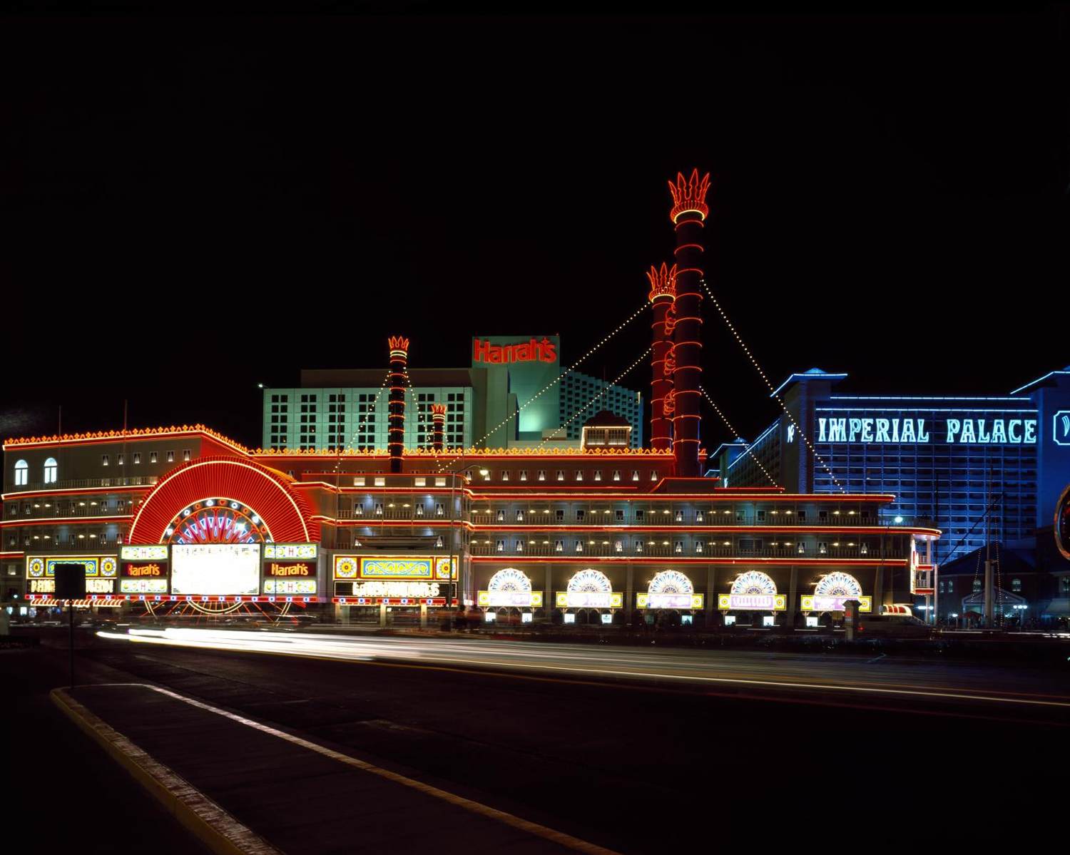Harrah's riverboat-themed facade and the Imperial Palace sign lit in neon at night with car light trails on the Strip