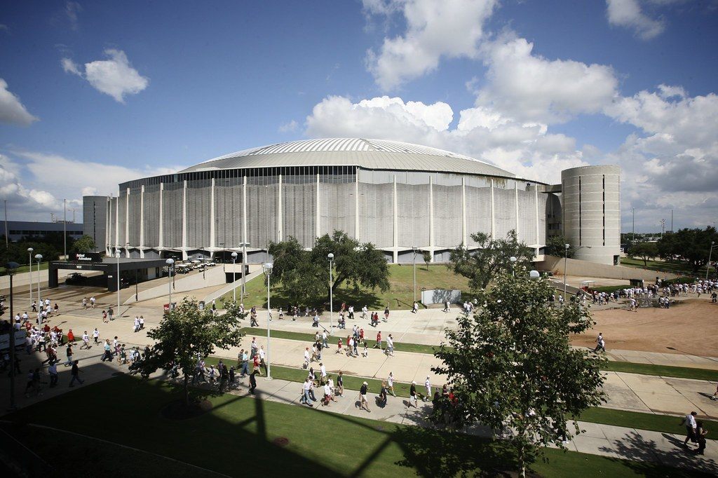 The Astrodome's ribbed white dome and round stair towers under a blue sky, with crowds walking along the paths of Reliant Park in front
