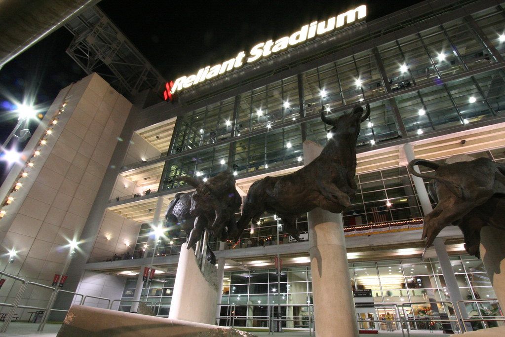 Bronze bull sculptures in front of the lit glass entrance of the stadium at night, with the Reliant Stadium sign glowing above