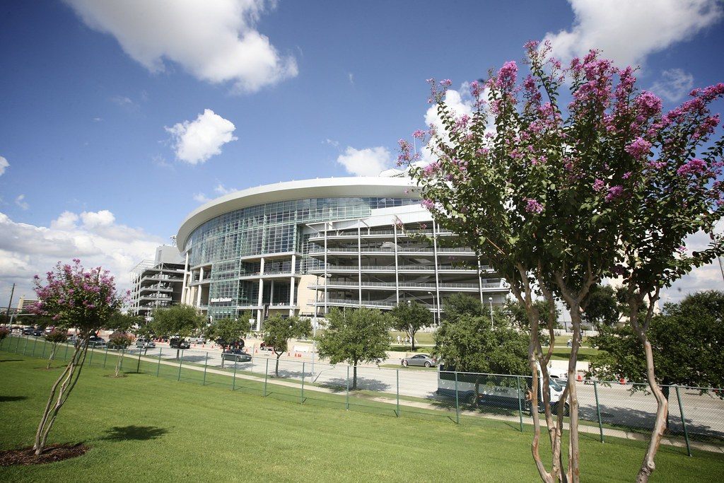 The curved glass front of the stadium in Reliant Park behind a lawn, trees and flowering crape myrtles, with cars on the park road below