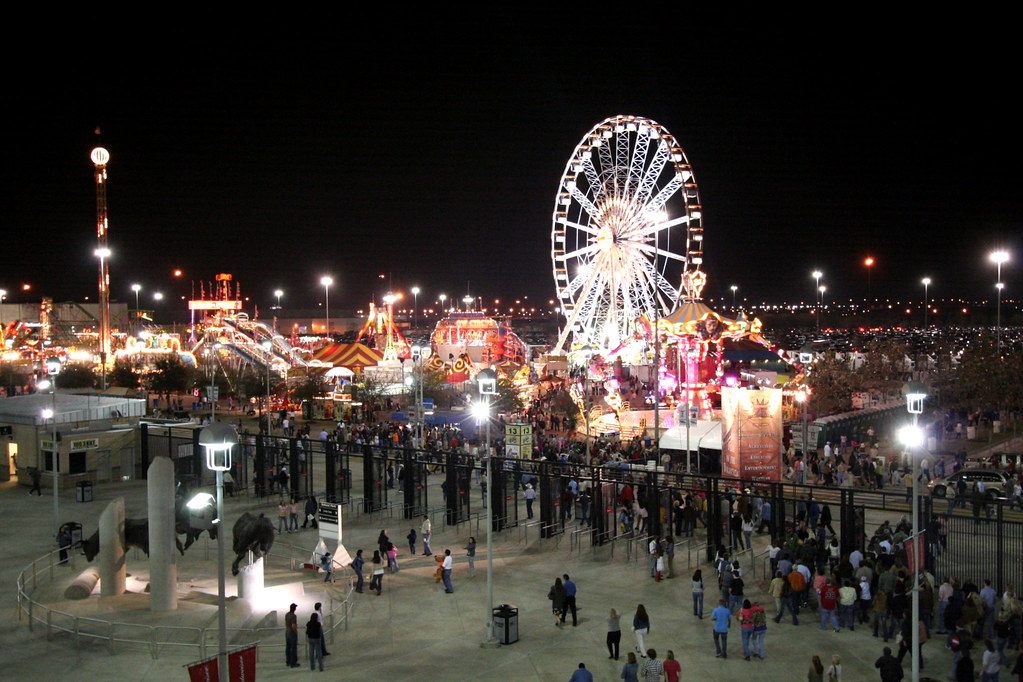 Visitors stream across the Reliant Park plaza at night toward the lit Ferris wheel and rides of the rodeo carnival