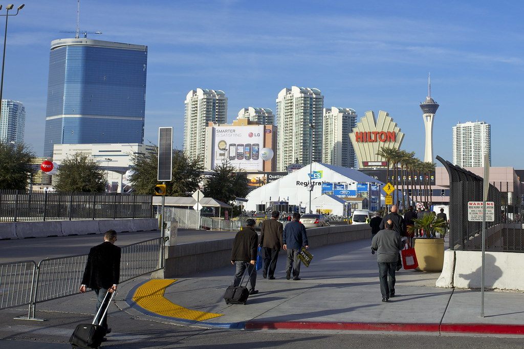 CES visitors with rolling bags walking along Paradise Road, with the tall blue-glass Fontainebleau tower and the Strat behind them