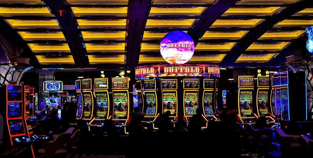Rows of lit slot machines under a glowing coffered ceiling and a spherical video display on the Resorts World casino floor