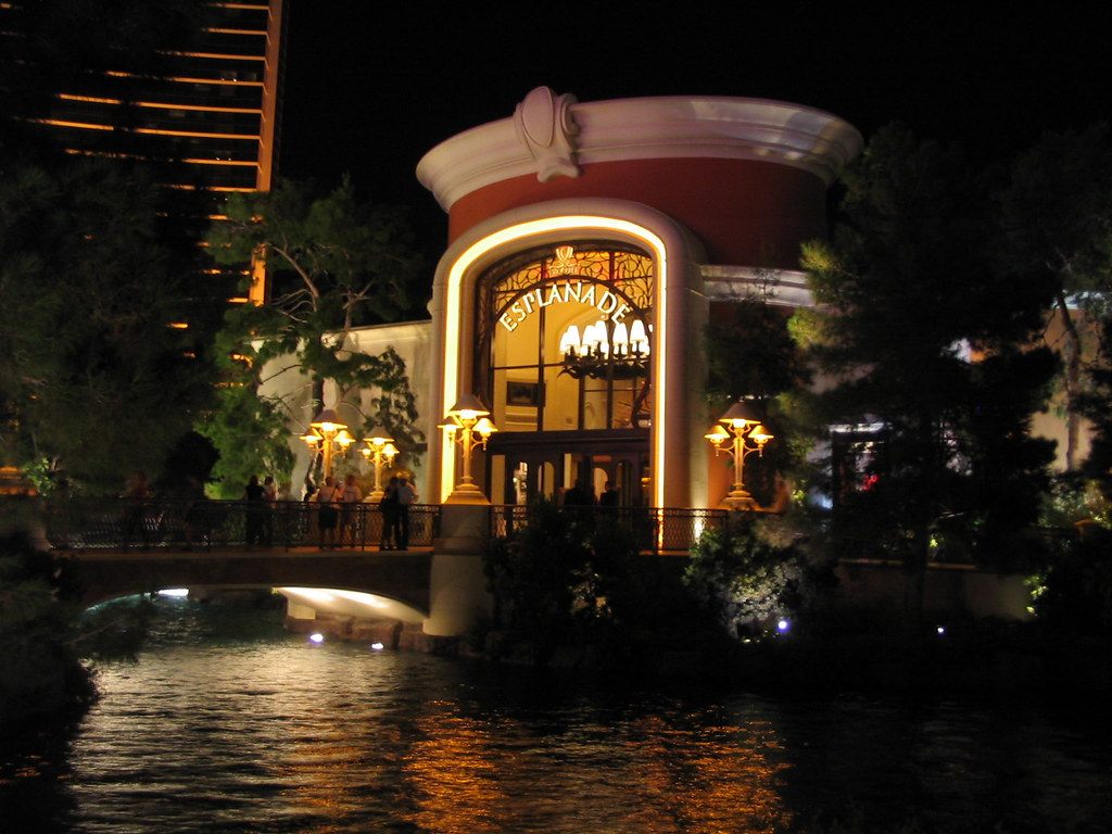 The lit entrance to the Esplanade shops at Wynn Las Vegas at night, beside a footbridge over a pond
