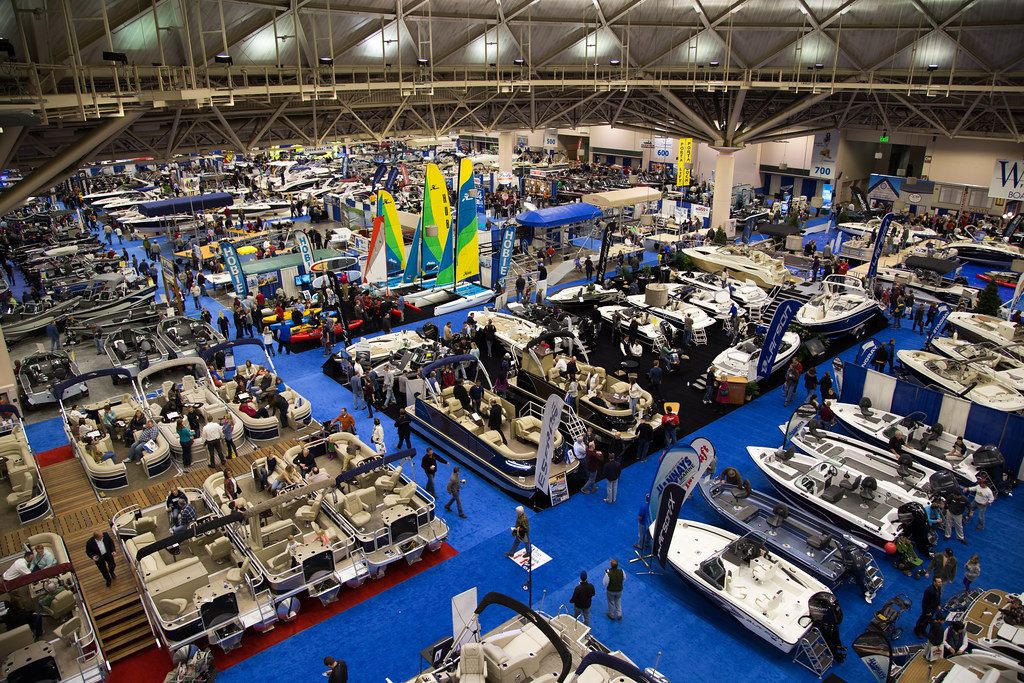 Rows of boats and pontoons on blue carpet at the Minneapolis Boat Show, seen from above under the exhibit hall's steel trusses