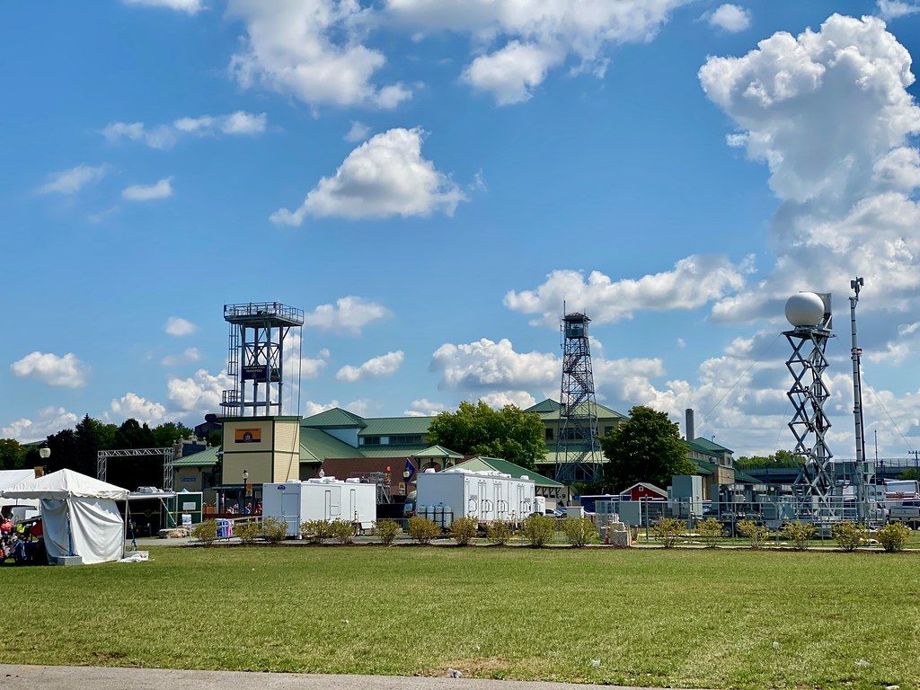 Green-roofed fair buildings, a lookout tower and lighting towers behind a lawn on the New York State Fairgrounds, with portable buildings lined up in front
