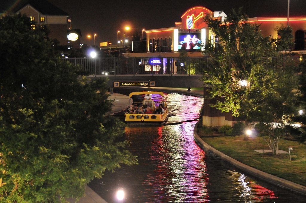 A lit water taxi cruising the Bricktown Canal at night past trees and restaurant signs