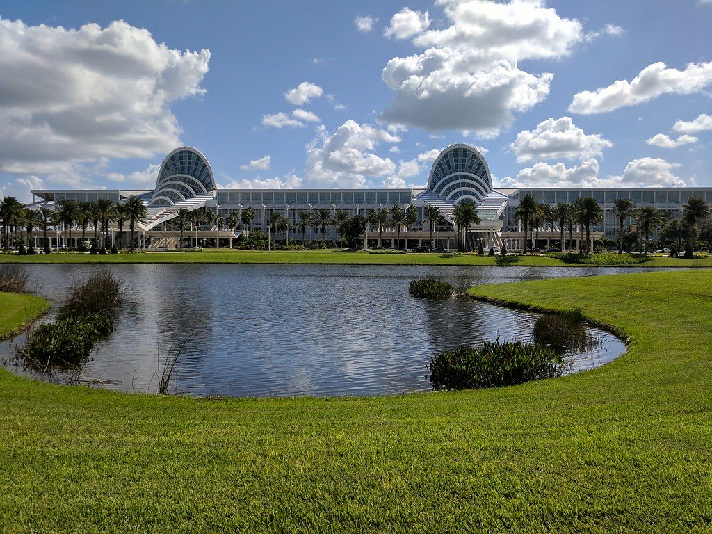 The Orange County Convention Center with its two glass fan-shaped entrance domes, seen across a pond and lawn under a cloudy blue sky