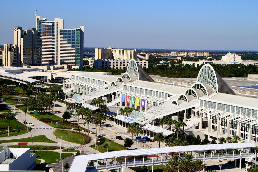Aerial view of the Orange County Convention Center West Building with its glass fan-shaped entrances, a covered walkway bridge in front and the hotel towers rising at left
