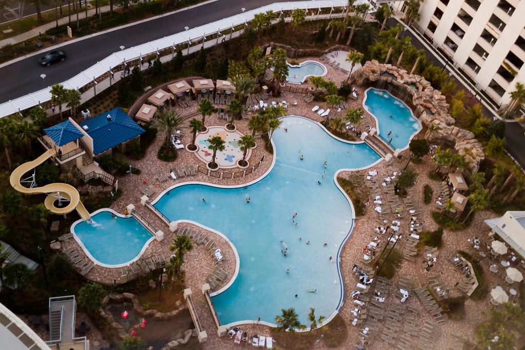 Aerial view of a large free-form resort pool with a water slide, cabanas, palm islands and rows of loungers beside a hotel tower