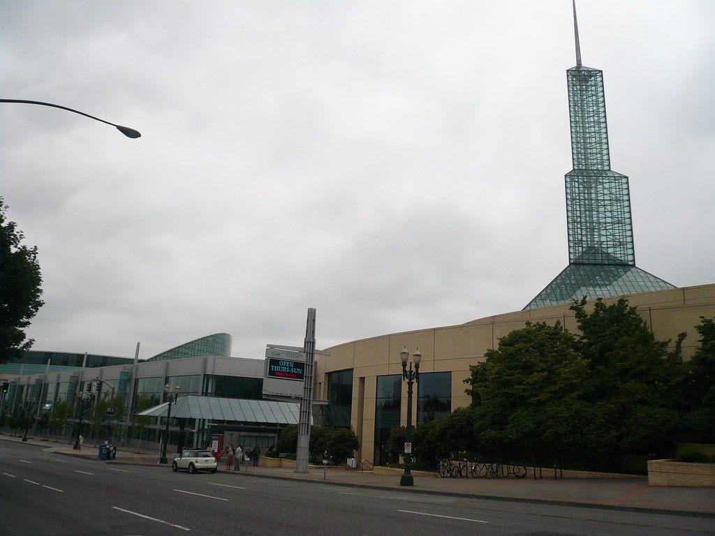 One glass spire above the long tan facade and glass entry canopy of the Oregon Convention Center on a cloudy day