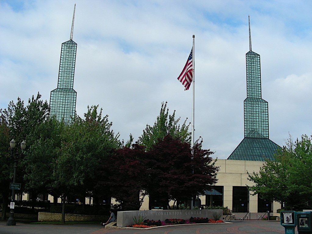 The two green glass spires of the Oregon Convention Center rising above trees and a flagpole at the main entrance