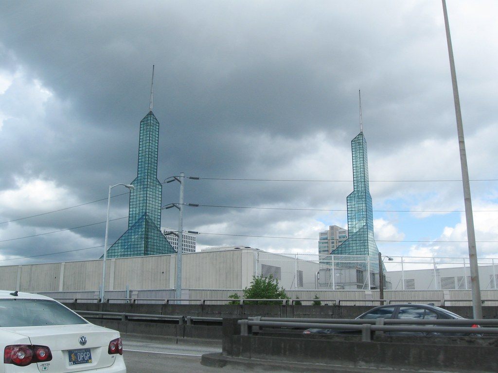 Both glass spires of the Oregon Convention Center seen over a concrete wall from a nearby road under a cloudy sky