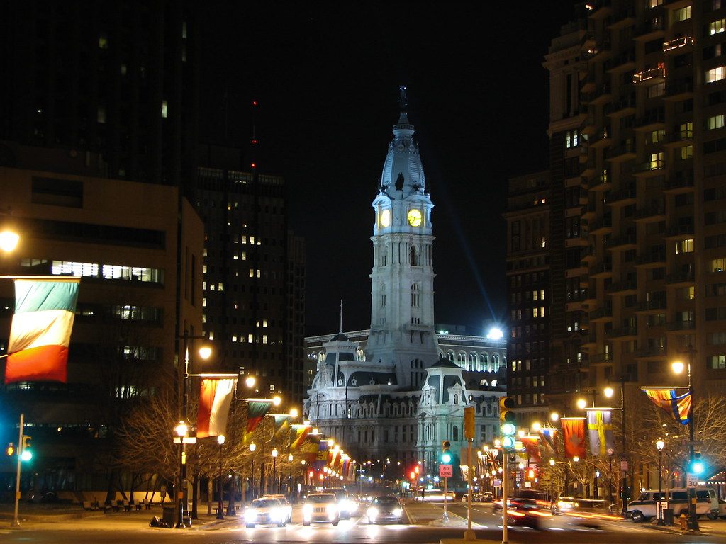 Philadelphia City Hall lit up at night at the end of a flag-lined boulevard, with traffic and towers on both sides