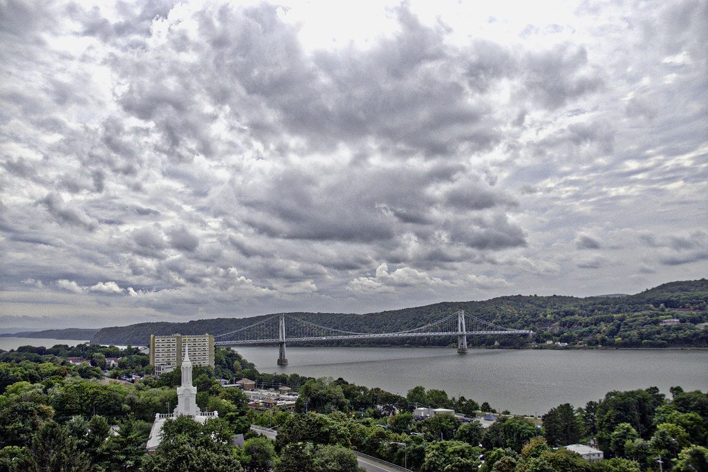 The Mid-Hudson Bridge spanning the Hudson River below a cloudy sky, with downtown Poughkeepsie and a ten-story hotel in the foreground